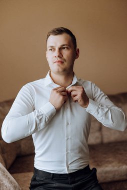 Beyaz gömlekli bir adam odanın penceresinin yanında durur ve yakasındaki ve kollarındaki düğmeleri bağlar. Hazır ol. Bir adamın şık bir iş portresi, yakın çekim fotoğrafı. Damat hazırlanıyor..