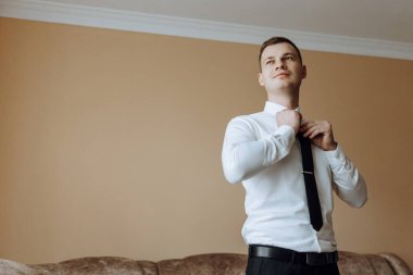 Beyaz gömlekli genç bir adam odanın penceresinin yanında duruyor ve kravat takıyor. Damat sabah giyinir ve düğün için hazırlanır..