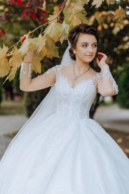 Düğün portresi. Saçında süs olan esmer bir gelin yüzüne yaprak tutarak poz veriyor. Sonbahar düğünü. Güzel makyaj ve saç. Kutlama için hazırlık.