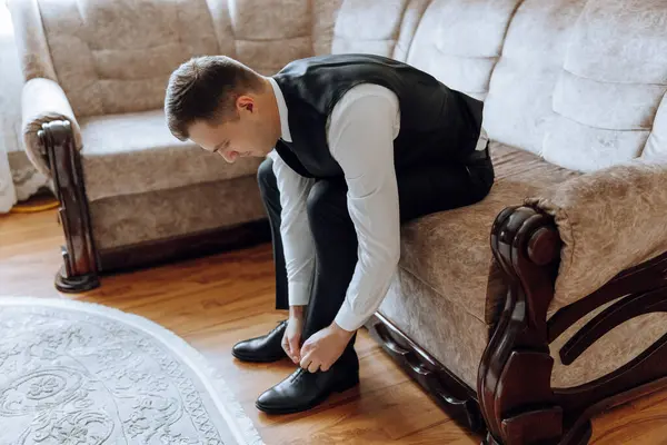 A man is putting on his shoes while sitting on a sofa in a hotel room. A young entrepreneur is preparing to go on a business trip. Businessman preparing for travel in home bedroom early in the morning
