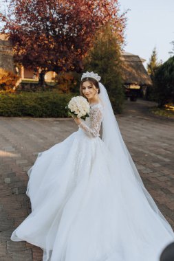 Uzun trenli beyaz elbiseli esmer bir gelin elbiseyi tutar ve taş patikadan aşağı doğru yürür. Sonbahar. Doğada düğün fotoğrafı çekimi. Güzel saç ve makyaj. kutlama