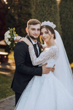 Şık, genç bir damat ve uzun beyaz bir elbise içinde güzel bir gelin ve ellerinde bir buket ile uzun bir duvak, sonbahar doğasında parkta sarılıyorlar. Yeni evlilerin düğün portresi..