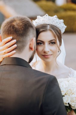 Elinde bir buket çiçekle kameraya bakarken kocasına sarılan gülümseyen bir gelinin portresi. Güzel bir düğün kutlaması. Duvağı olan düğün saç stili. Birbirine aşık çift.