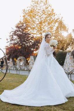 Düğün fotoğrafı. Dantelli, dolgun elbiseli esmer bir gelin elinde bir buket tutarak bahçede yürüyor, içtenlikle gülümsüyor. Harika saç ve makyaj. Moda ve tarz. Kutlama.