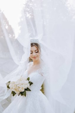 Düğün portresi. Dantelli bir elbise ve şık bir taç giymiş esmer bir gelin, uzun bir peçeyle örtülü bir buket tutarak doğada poz veriyor. Güzel makyaj. Sonbahar fotoğraf çekimi