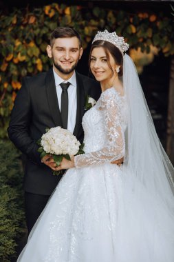 Şık, genç bir damat ve uzun beyaz bir elbise içinde güzel bir gelin ve ellerinde bir buket ile uzun bir duvak, sonbahar doğasında parkta sarılıyorlar. Yeni evlilerin düğün portresi..