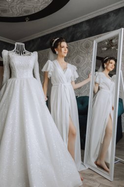 Sabahleyin, inanılmaz güzel ve lüks bir gelin, beyaz bir bornoz giymiş, yatak odasındaki aynanın önünde poz veriyor. Gelinin şaşaalı saç stili ve doğal makyajlı sabahı..