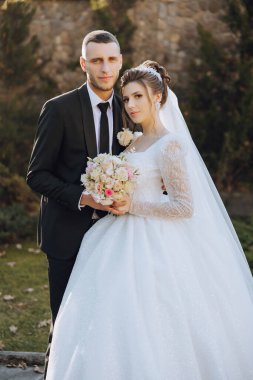 Şık, genç bir damat ve uzun beyaz bir elbise içinde güzel bir gelin ve ellerinde bir buket ile uzun bir duvak, sonbahar doğasında parkta sarılıyorlar. Yeni evlilerin düğün portresi..