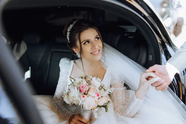 A beautiful bride, sitting in a car, gives her hand to her husband. A beautiful bride with a bouquet of flowers in her hands is sitting in a stylish expensive car.