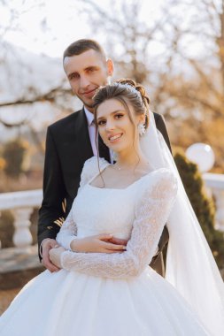 Şık, genç bir damat ve uzun beyaz bir elbise içinde güzel bir gelin ve ellerinde bir buket ile uzun bir duvak, sonbahar doğasında parkta sarılıyorlar. Yeni evlilerin düğün portresi..