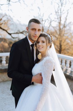 Şık, genç bir damat ve uzun beyaz bir elbise içinde güzel bir gelin ve ellerinde bir buket ile uzun bir duvak, sonbahar doğasında parkta sarılıyorlar. Yeni evlilerin düğün portresi..