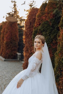 Düğün portresi. Sarışın gelin, dantelli, omuzları açık, ağaçların arka planında poz veriyor. Güzel saç ve makyaj. Sonbahar. Gün ışığı. kutlama.
