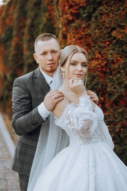 Şık, genç bir damat ve uzun beyaz bir elbise içinde güzel bir gelin ve ellerinde bir buket ile uzun bir duvak, sonbahar doğasında parkta sarılıyorlar. Yeni evlilerin düğün portresi..