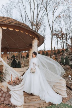 Şehvetli beyaz elbiseli esmer gelin elinde buket, poz veriyor. Duvak havaya fırlatıldı. Güzel saç ve makyaj. Doğada düğün fotoğrafı çekimi. Elbisenin uzun treni.
