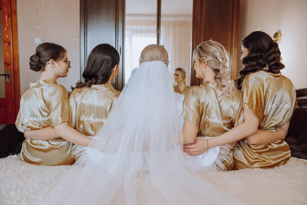 A beautiful bride and her bridesmaids are sitting in a room on a bed with their backs turned in identical robes with the inscription bridesmaid. Wedding celebration. Happy girls.
