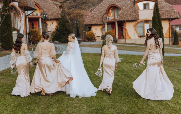 Wedding photography. A blonde bride in a long wedding dress and her friends in beige dresses walk to the houses, rear view. Autumn wedding. Young girls. Posing