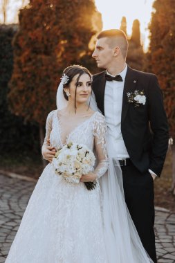 Şık, genç bir damat ve uzun beyaz bir elbise içinde güzel bir gelin ve ellerinde bir buket ile uzun bir duvak, sonbahar doğasında parkta sarılıyorlar. Yeni evlilerin düğün portresi..