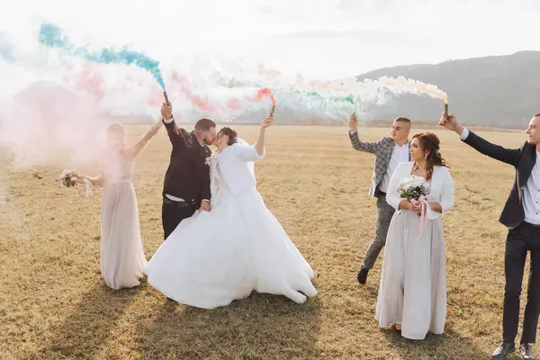 Wedding photo session in nature. Bride and groom and their friends in a field having fun holding colored smoke. Bright colors. Autumn wedding. Friendship. A group of young people. Celebration