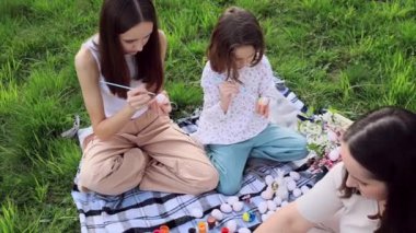 Farklı yaşlarda ve Avrupa görünümlü üç kızdan oluşan bir aile yeşil çimlerin üzerinde bahçede oturuyor ve Paskalya yumurtalarını boyamakla meşguller. Paskalya gelenekleri. Bahar tatili.