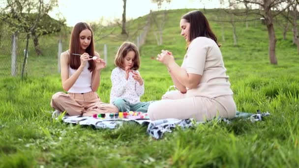 一个由三个不同年龄和长相的欧洲姑娘组成的家庭正坐在绿草上的花园里 正在画复活节彩蛋 复活节传统 — 图库视频影像
