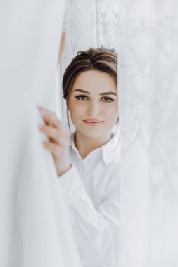 Perdenin önünde duran bir kadın, perdeden dışarı bakıyor. Beyaz bir gömlek giyiyor ve saçları topuz şeklinde. Merak ve entrika kavramı