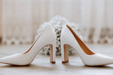 Ayak parmağında yüzük olan iki beyaz topuklu. Ayakkabılar ahşap bir zemine, bir buket çiçeğin yanına yerleştirilir. Hem ayakkabılar hem de çiçekler klasik olduğu için zarafet ve entelektüellik kavramı