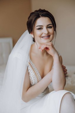 Kafasında beyaz bir peçe olan bir kadın yatakta oturuyor. Gülümsüyor ve elleri dizlerinin üstünde.