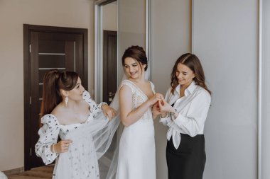 Üç kadın düğün için hazırlanıyor. Biri beyaz bir elbise giyiyor ve diğer ikisi de elbisesine yardım ediyor.