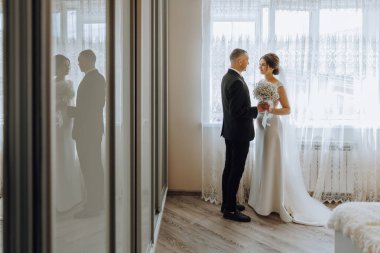 Gelin ve damat, gelin elinde buketle pencerenin önünde dururlar. Aşk ve mutluluk kavramı, çift birlikte yeni bir hayata hazırlanırken