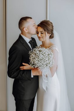 Bir gelin ve damat birbirlerine sarılıp öpüşüyorlar. Gelin elinde bir buket beyaz çiçek tutuyor.