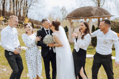 Bir grup insan, ortasında bir gelin ve damat olan bir resim için poz veriyor. Gelin bir buket tutuyor ve damat onu öpüyor. Gruptaki diğer insanlar gülümsüyor.