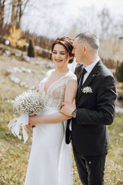 Bir gelin ve damat tarlada fotoğraf çektirmek için poz veriyorlar. Gelin elinde bir buket beyaz çiçek tutuyor ve damat takım elbise giyiyor. Sahne mutlu ve romantik.