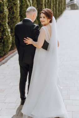 Bir gelin ve damat, tuğla kaldırımda fotoğraf çektirmek için poz veriyor. Gelin beyaz bir elbise giyiyor ve damat siyah bir takım elbise giyiyor. İkisi de gülümsüyor ve birlikte olmaktan mutlu görünüyorlar.