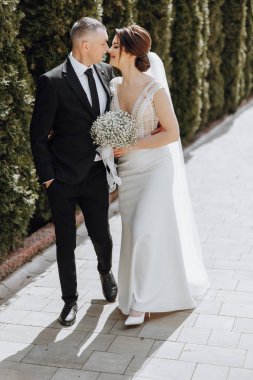 Bir adam ve bir kadın kaldırımda yürüyorlar, adam takım elbise ve kravat takıyor. Kadın elinde bir buket çiçek tutuyor.