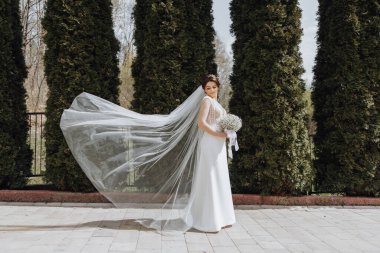 Beyaz elbiseli bir kadın çitin önünde duruyor. Elinde bir buket çiçek ve duvak tutuyor.