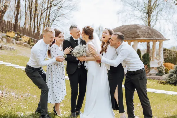 A group of people are posing for a picture, including a bride and groom. The bride is holding a bouquet of flowers, and the groom is smiling. The group is standing in a grassy area
