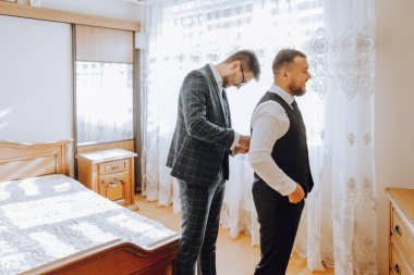 İki adam yatak odasında giyiniyor. Bir adam takım elbise ve kravat takarken diğeri yelek giyiyor.