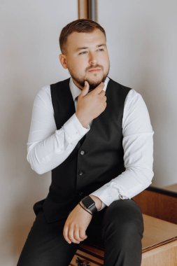 Siyah yelek ve beyaz gömlekli bir adam masanın üzerinde oturuyor. Saat takıyor ve sakalı var.