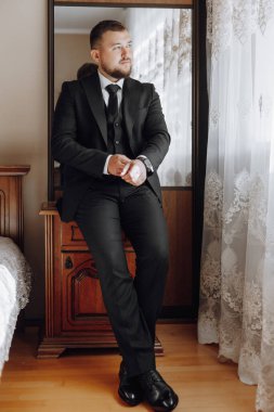 Takım elbiseli ve kravatlı bir adam aynanın önündeki şifoniyere oturur. O siyah bir takım ve beyaz bir gömlek giyiyor.