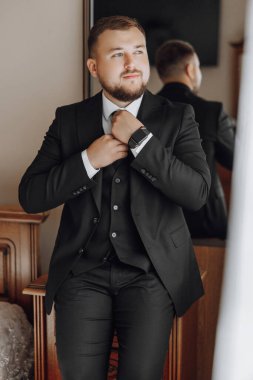 Takım elbiseli bir adam kravatını bağlamaya hazırlanıyor. Siyah beyaz bir saat ve kravat takıyor.