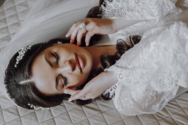 Bir kadın elleri yüzü üzerinde bir yatakta uzanıyor. Gelinlik ve yüzük takıyor. Kadın rahat bir konumda olduğu için samimiyet ve rahatlama kavramı.
