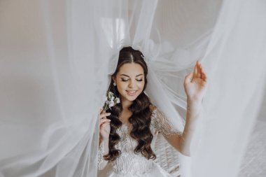 Saçında çiçekle beyaz bir peçenin altında oturan bir gelin. Gülümsüyor ve kameraya bakıyor.