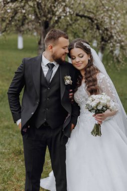 Siyah takım elbiseli damat beyaz elbiseli gelinini kucaklıyor. Düğün baharda. Doğada güzel bir çift