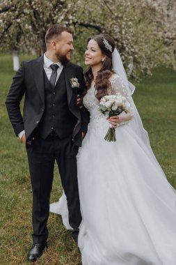 Siyah takım elbiseli damat beyaz elbiseli gelinini kucaklıyor. Düğün baharda. Doğada güzel bir çift