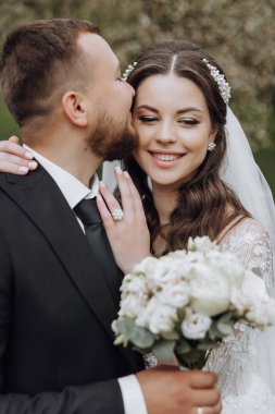 Bir gelin ve damat birbirlerinin dudaklarını öpüyor. Gelin elinde bir buket çiçek tutuyor.
