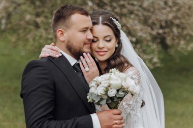 Gelin ve damat birbirlerine sarılıyorlar, gelin de bir buket çiçek tutuyor. Aşk ve mutluluk kavramı, çiftler düğün günlerini kutlarken