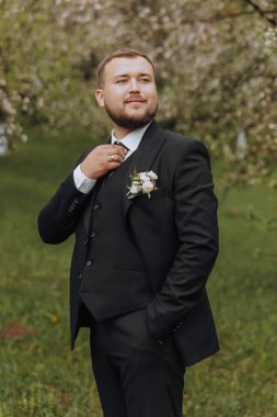 Siyah takım elbiseli damat doğadaki yeşil çimlerin üzerinde yürüyor. Klasik siyah takım elbiseli zarif bir adam. Bir adamın portresi.
