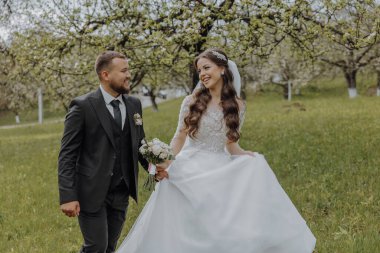 Damat ve gelini bahar bahçesindeki yeşil çimlerin üzerinde yürüyorlar. Gelin şık beyaz bir elbise içinde, damat siyah bir takım elbise içinde.