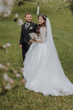 Damat ve gelini bahar bahçesindeki yeşil çimlerin üzerinde yürüyorlar. Gelin şık beyaz bir elbise içinde, damat siyah bir takım elbise içinde.