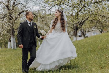 Damat ve gelini bahar bahçesindeki yeşil çimlerin üzerinde yürüyorlar. Gelin şık beyaz bir elbise içinde, damat siyah bir takım elbise içinde.